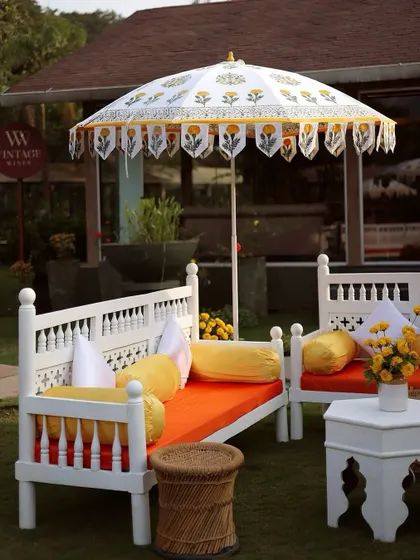 A vibrant Haldi seating area with traditional white wooden benches, colorful orange and yellow cushions, and custom-printed umbrellas.