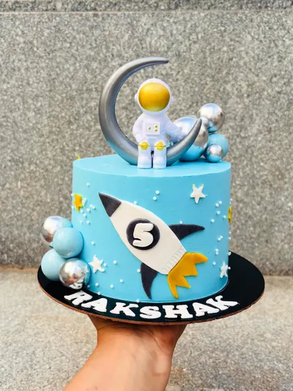 An out-of-this-world space theme cake. This design features an astronaut, a rocket ship, and planets, all on a vibrant blue buttercream galaxy.