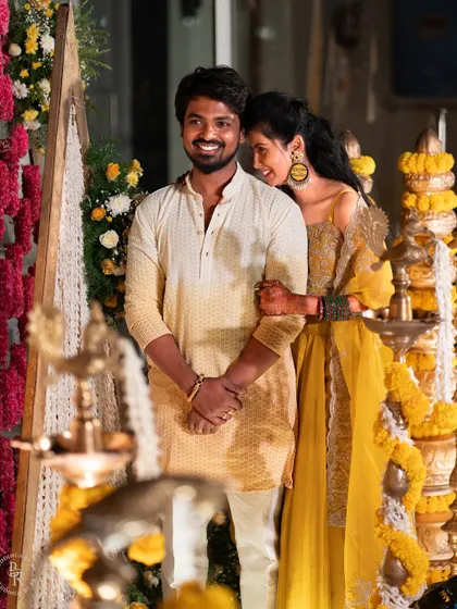 The couple shares a sweet, candid moment, framed by traditional brass lamps and floral decor at their Haldi.