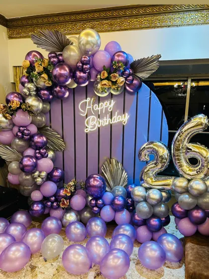 A modern purple paneled backdrop decorated with a lush arrangement of purple and silver balloons and dried palm leaves for a 26th birthday.