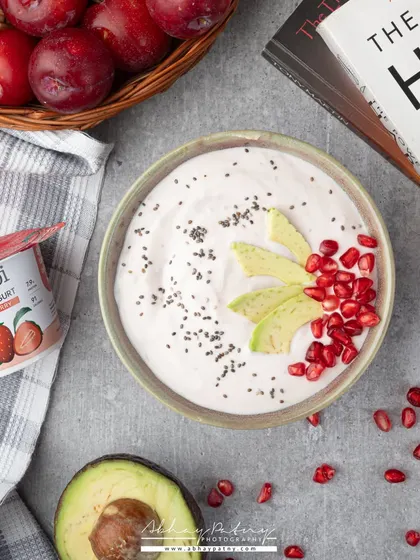This lifestyle shot shows Greek yogurt in a real-life breakfast setting. The composition with fresh fruits, a book, and natural light creates an aspirational and healthy image for the brand.