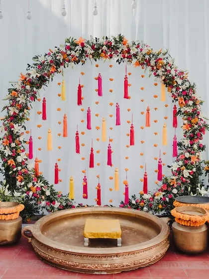 A beautiful circular floral backdrop for a Haldi ceremony, featuring a ring of colorful flowers and hanging tassels.