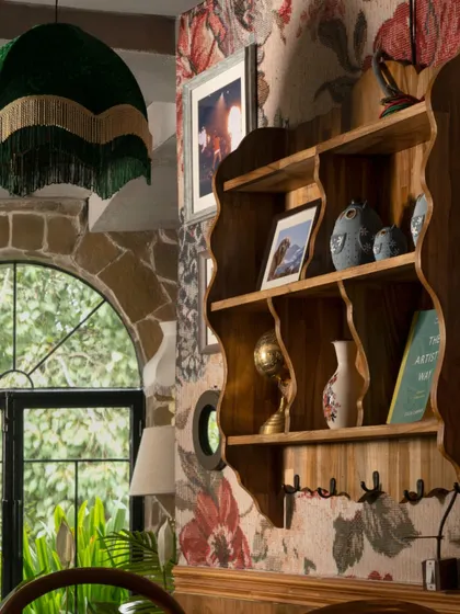 A scalloped wooden shelf holds a collection of curiosities, set against a backdrop of floral and stone textures. The fringed green lampshade adds a touch of playful maximalism to this eclectic corner.