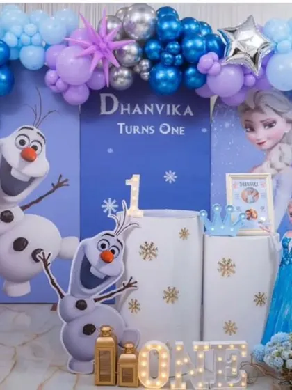 An elaborate Frozen theme setup for a first birthday, featuring a detailed backdrop with Elsa and Olaf, a stunning balloon arch with snowflake accents, and a light-up 'ONE' sign.