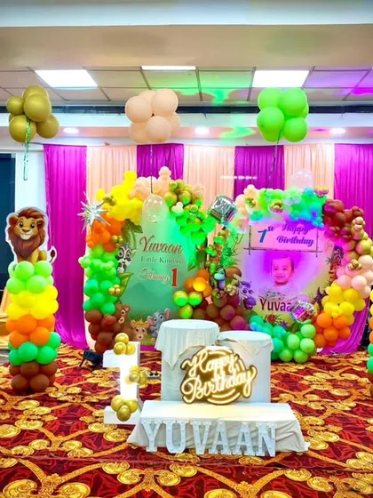 A vibrant jungle-themed stage for Yuvaan's first birthday. The decoration is filled with colorful balloons, various animal cutouts, and a custom "Happy Birthday" sign.