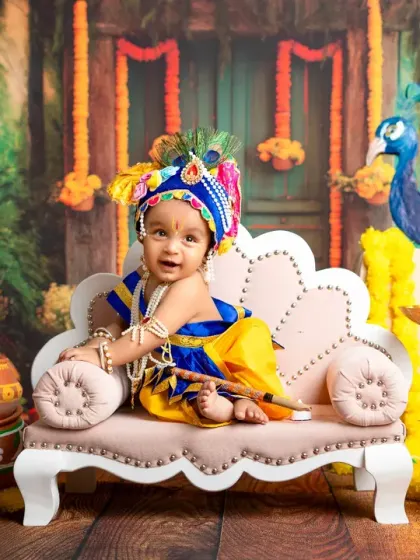 A beautiful, centered portrait of a baby in a full Krishna costume, surrounded by a vibrant and detailed set. This is a perfect example of a fully styled themed shoot.
