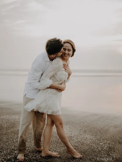 A candid and joyful pre-wedding moment on the beach, with the groom kissing the bride's cheek as she laughs.