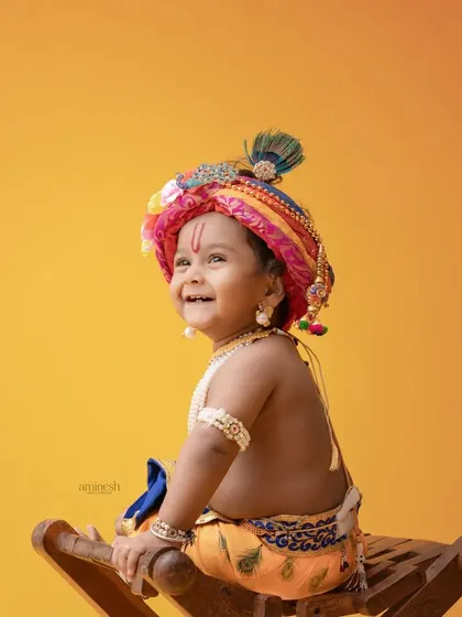 A bright and cheerful close-up of a baby in a Krishna costume against a vibrant yellow background. The focus is on the child's infectious smile and colorful turban.