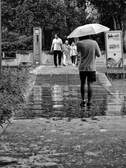 A black and white shot from behind a man walking on a wet path with an umbrella. The reflections on the ground and the sense of solitary movement define this monsoon scene.