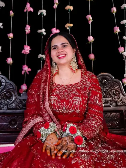The bride, seated and smiling, in her magnificent red reception outfit. The floral backdrop and her joyful expression create a picture of pure celebration.