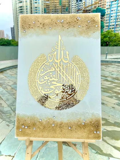 A vertical shot of the white and gold Ayat al-Kursi piece on an easel against a modern city backdrop.