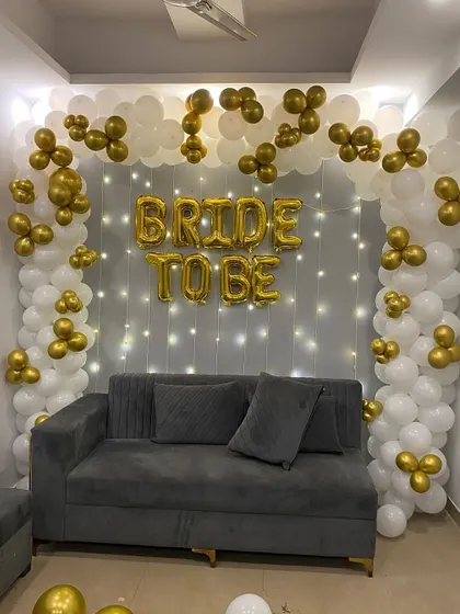 A close-up of the white and gold balloon arch for a 'Bride to Be' party. The fairy light curtain behind the balloons adds a warm and sparkling glow to the entire setup.