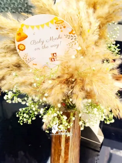 A beautiful centerpiece from the rustic baby shower, featuring pampas grass and baby's breath in a vase, with a custom 'Baby Mushi on the way!' sign.