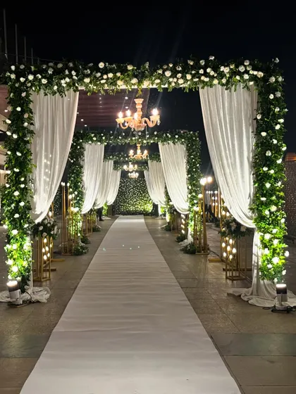 A majestic entrance tunnel for a Walima or reception at Crowne Plaza. The walkway is lined with arches of white drapes and lush green and white florals, lit by elegant chandeliers.