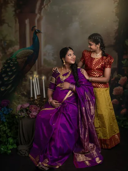 An expecting mother sits gracefully while her daughter stands beside her, both dressed in traditional Indian clothing. The scene is set against a beautiful painted backdrop, creating a regal and timeless portrait.