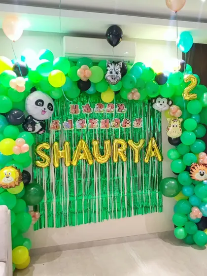 A lush jungle-themed birthday decoration for a two-year-old. I created a full balloon arch in shades of green to mimic a jungle canopy, accented with cute animal foil balloons like a panda, zebra, and lion. The green shimmer backdrop and gold foil name "SHAURYA" make this a fun and adventurous photo corner.