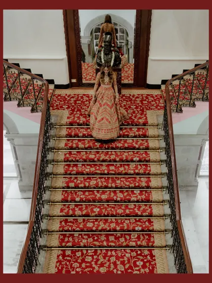 A high-angle shot of the bride and her entourage on the grand red-carpeted staircase. The symmetry and rich colors create a visually stunning and opulent image.