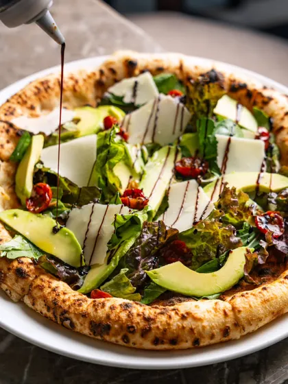 Balsamic glaze being drizzled over a fresh salad pizza. The motion of the drizzle adds a layer of visual interest to the photograph.