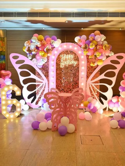 A 6th birthday celebration with a butterfly theme. The decor features large marquee number '6', pink butterfly wings, and balloon columns against a wooden wall backdrop.