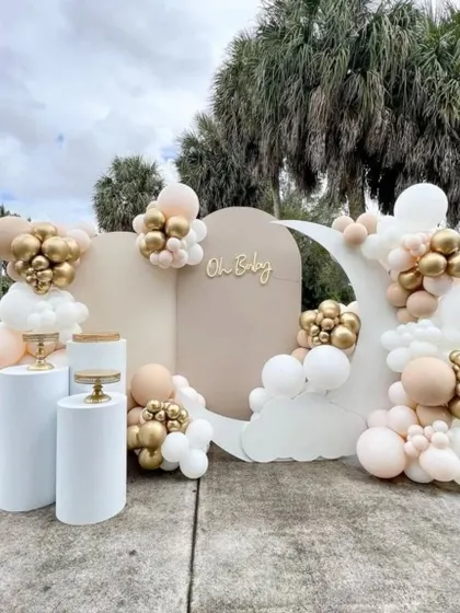 A chic and minimalist outdoor baby shower setup. This design uses neutral backdrops, a crescent moon prop, and a simple balloon arrangement in white, beige, and gold for an understated, elegant look.