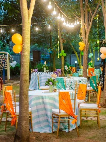 An outdoor seating area for a Moana-themed party. The tables are set up under trees strung with lights, and the colorful chair sashes add a festive, tropical touch.