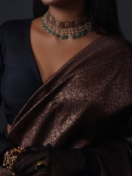 A close-up on the jewelry and fabric from the 'Raaga' series. This shot emphasizes the beautiful contrast between the dark, shimmering saree, the emerald green choker, and the delicate sheer gloves.