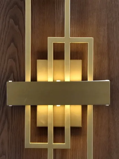 A close-up view of the fine details that elevate our designs. This image showcases a custom-designed geometric wall sconce with a brushed gold finish, mounted on a rich wood veneer panel, adding a touch of artistic flair.