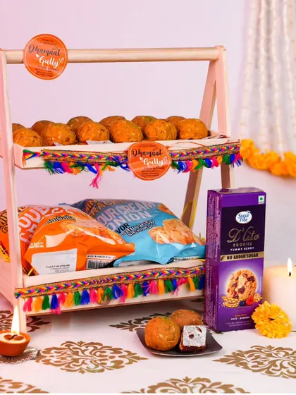 This fun and casual Diwali hamper is perfect for snack lovers. It features a two-tiered stand loaded with packets of chips, cookies, and my homemade mathris, all presented with festive diyas.