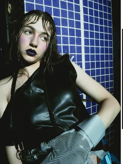 A close-up portrait with dramatic, dark makeup against a blue tiled wall. The wet-look hair and intense gaze add to the raw, edgy feel of this experimental shoot.