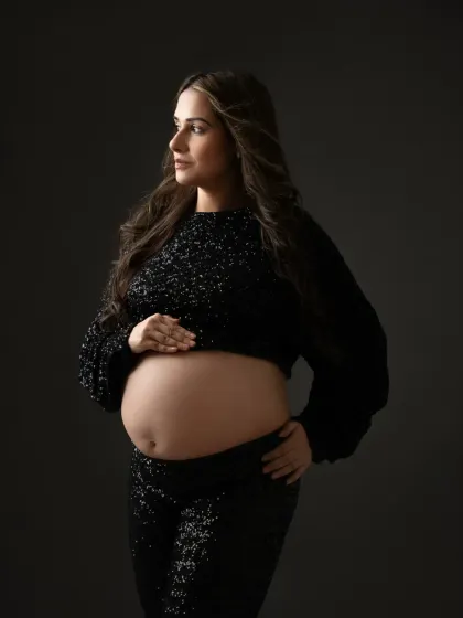 A thoughtful, powerful pose showing off the baby bump. The dark, moody lighting complements the sparkling outfit for a sophisticated and modern maternity photo.