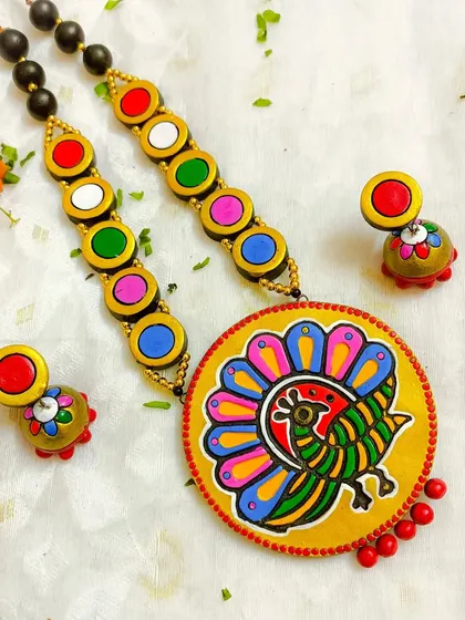 This terracotta necklace features a block-printed peacock motif, combining printmaking techniques with hand-painting for a vibrant, layered look. The piece is finished with colorful beads and matching jhumkas.
