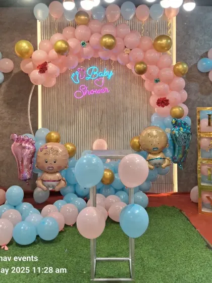 A front view of a pink and blue themed baby shower decoration. It includes a balloon arch on a ring stand, a neon sign, baby-shaped foil balloons, and 'BABY' blocks.