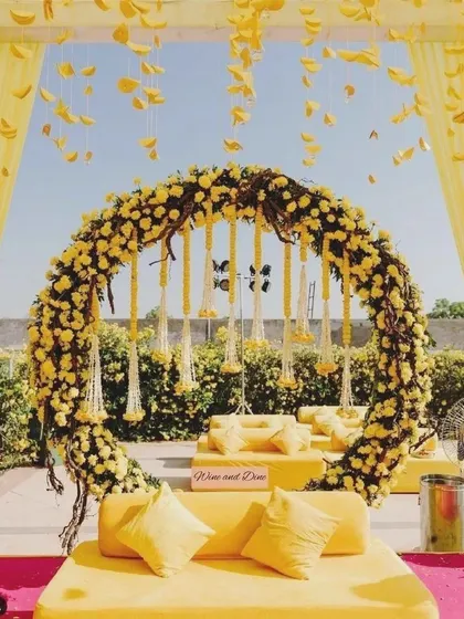 A beautiful outdoor Haldi setup with a circular arch made of yellow flowers and hanging jasmine strings. The all-yellow seating arrangement creates a sunny and cheerful vibe.
