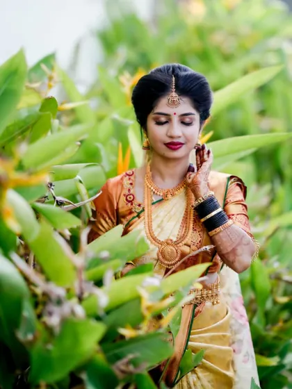 Surrounded by nature, this bride's beauty shines through. The makeup features a classic red lip and defined eyes, which stand out beautifully against her cream and gold saree.