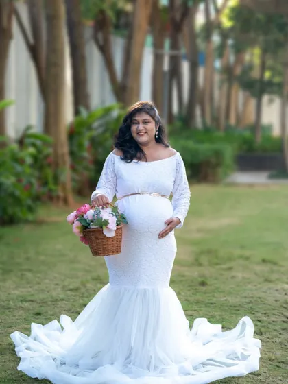 A lovely outdoor shot of a mom-to-be in a white lace gown, holding a basket of flowers.