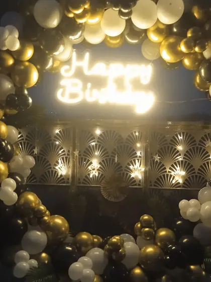 A nighttime birthday party setup featuring a circular balloon arch in gold, black, and white. The 'Happy Birthday' neon sign glows in the center, creating a perfect focal point for an evening celebration.