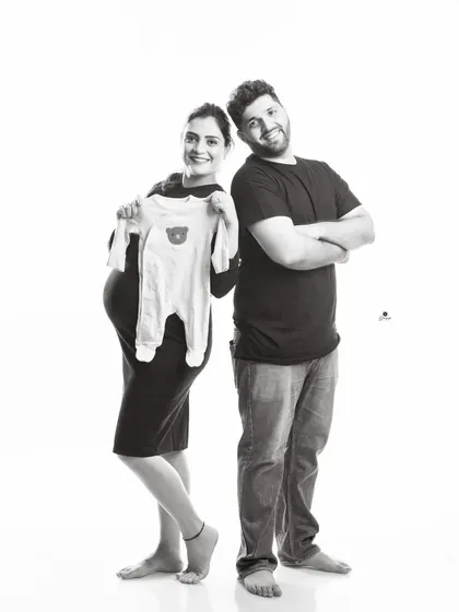 A fun and casual black and white portrait. The couple stands back-to-back, with the mom-to-be proudly holding up a baby onesie, capturing their happy and relaxed dynamic.