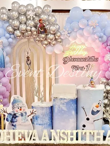 A full stage setup for a Frozen-themed first birthday. The decor includes character cutouts of Elsa, Anna, and Olaf, a "Happy Birthday" neon sign, and a beautiful balloon arch with silver snowflake accents.