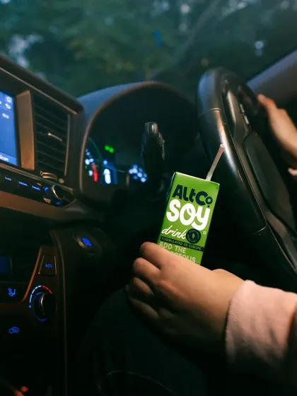 A moody lifestyle shot of Alt Co.'s soy drink held by a person in a car at night. The ambient light from the dashboard creates an intimate, on-the-go moment.