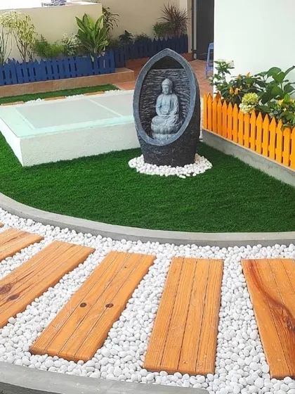 Add a touch of tranquility to your garden. This design incorporates a serene Buddha water feature as a focal point, surrounded by artificial grass, pebble pathways, and colorful fencing.