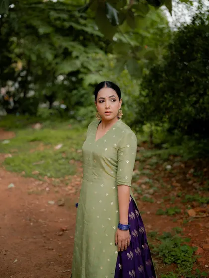A full-length portrait of actress Anupama Gowda in a traditional outfit, standing in a natural, rustic setting. The composition is simple and elegant, focusing on her graceful presence.