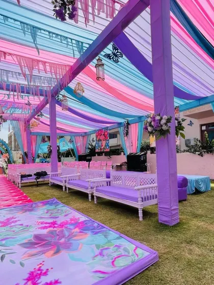 An outdoor Haldi setup featuring a pastel theme with lavender, pink, and blue drapes creating a beautiful canopy. The seating area is designed with matching purple couches for a cohesive look.