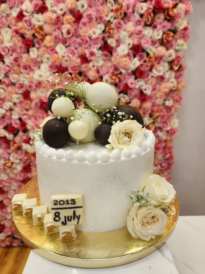 An elegant white anniversary cake decorated with chocolate spheres, fresh white roses, and a custom date marker.