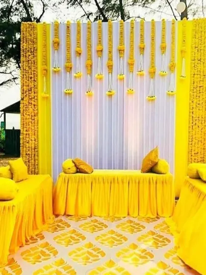 This haldi decor features a clean, modern aesthetic with a backdrop of white and yellow panels. The hanging floral strings add a traditional touch, while the symmetrical seating arrangement provides a neat and organized look.