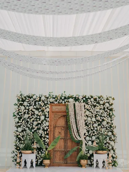 A beautiful and minimalist backdrop for a Haldi ceremony. A wall of white flowers provides a clean and elegant setting, with a traditional wooden door adding a touch of rustic charm.