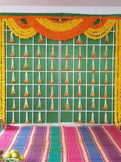 A traditional Haldi backdrop with a green base, decorated with hanging flower strings and a large toran of orange and yellow marigolds.