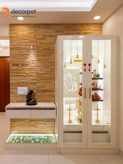 This pooja unit combines a stone-clad wall with a clean, white cabinet. The glass doors are adorned with hanging bells, and the floor-level glass enclosure with pebbles adds a unique, natural element.