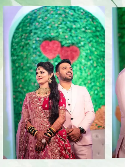 A classic reception stage photo. The couple stands together, looking elegant in their reception outfits against a backdrop of green leaves and red hearts.