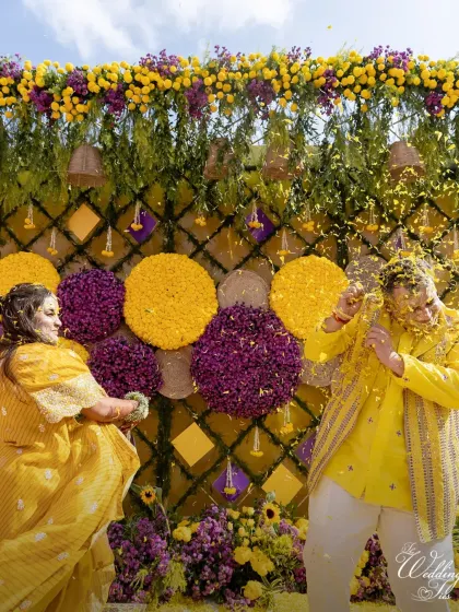 A playful moment between the couple during their Haldi, set against a stunning floral wall of yellow and purple, adding a unique twist to the traditional decor.