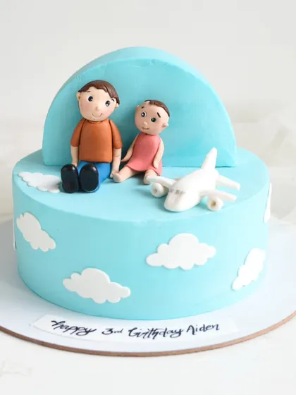 A sweet travel-themed cake featuring handmade fondant figures of a father and child sitting among the clouds with an airplane. This cake has two delicious tiers of milk chocolate and black forest.